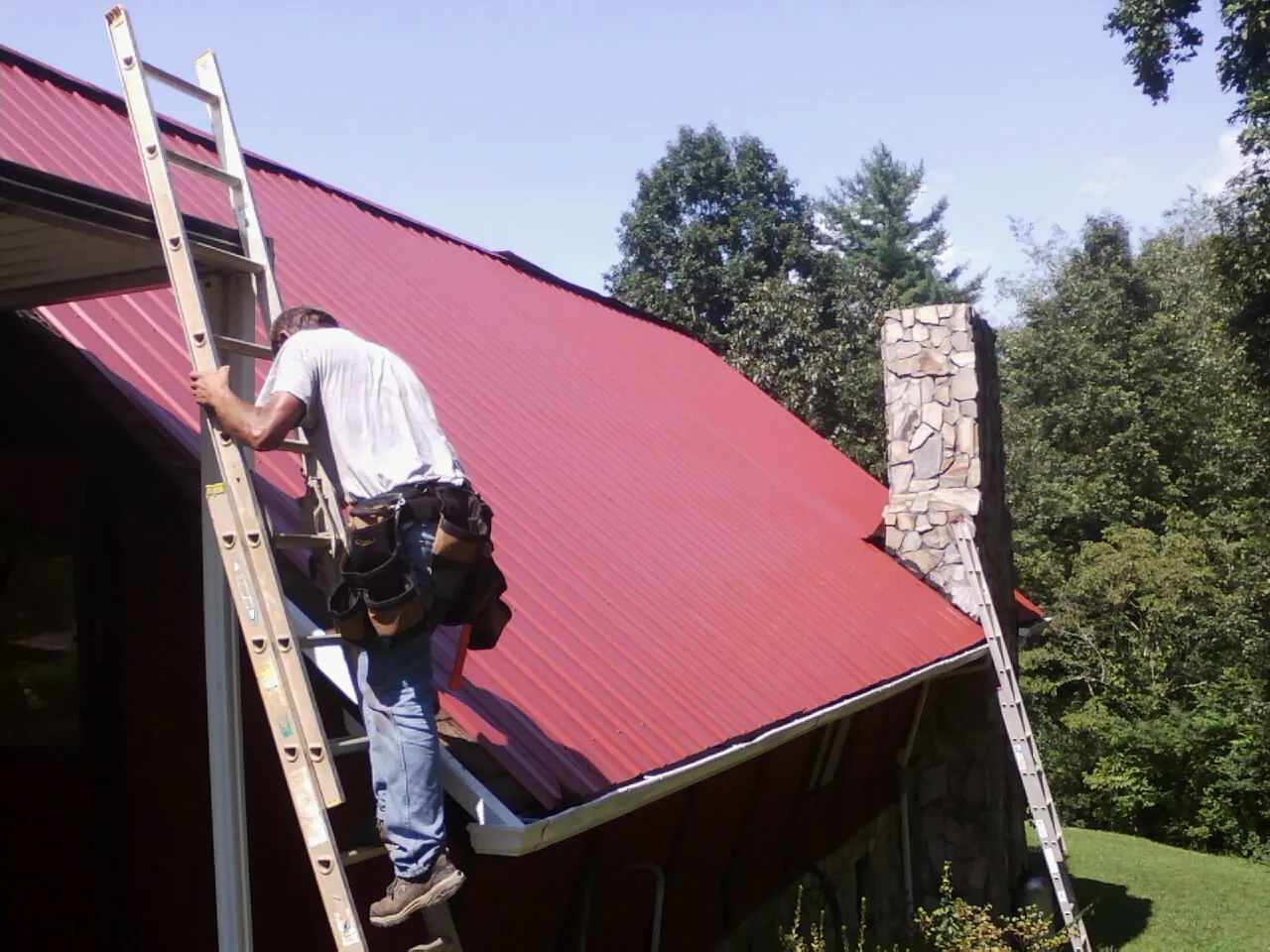 Roofer on a ladder beside a red metal roof and stone chimney — Soffit & Fascia Repair by Roofing Brothers in Shasta Lake